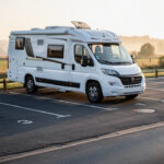 Où stationner un camping-car en France en toute légalité ? Camping-car blanc stationné discrètement sur une aire dédiée au lever du soleil, avec des emplacements et des repères de stationnement visibles.