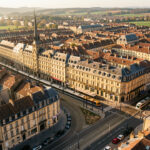 Quels quartiers choisir pour acheter à Dijon selon les prix au m² en 2026 ? Vue aérienne réaliste de Dijon montrant le centre historique, les toits bourguignons et les lignes de tramway baignés par la lumière dorée de fin d’après-midi.